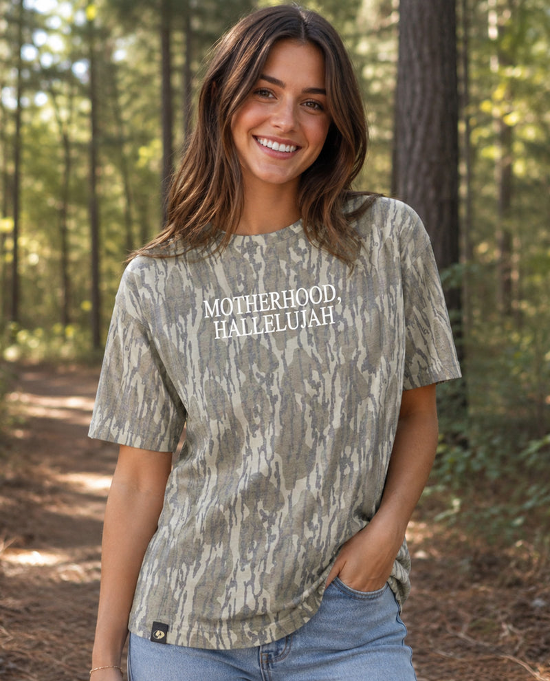 A woman smiling outdoors in a forest wearing a short-sleeve t-shirt with a brown and beige tree-bark camouflage pattern and the words 'MOTHERHOOD Hallelujah' printed in white across the chest.