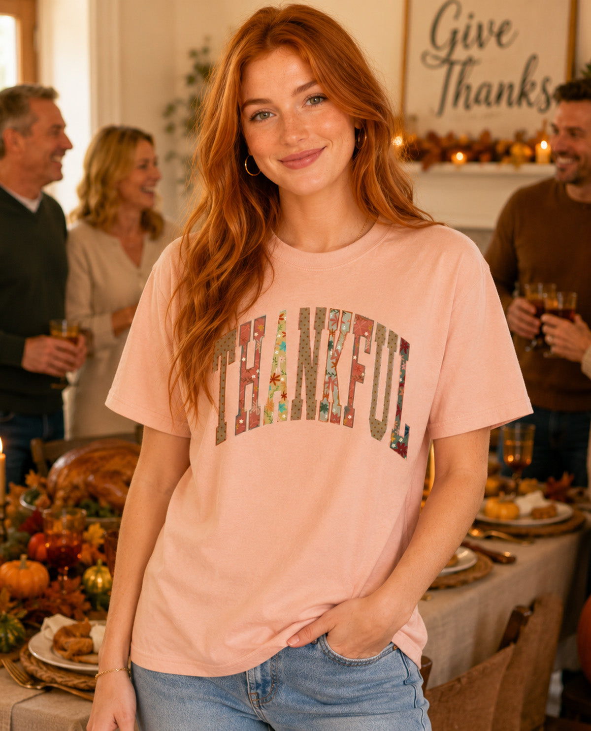 A woman wearing a light peach-pink t-shirt with 'THANKFUL' printed in a floral autumn-themed font, standing in a cozy, decorated Thanksgiving dining room.