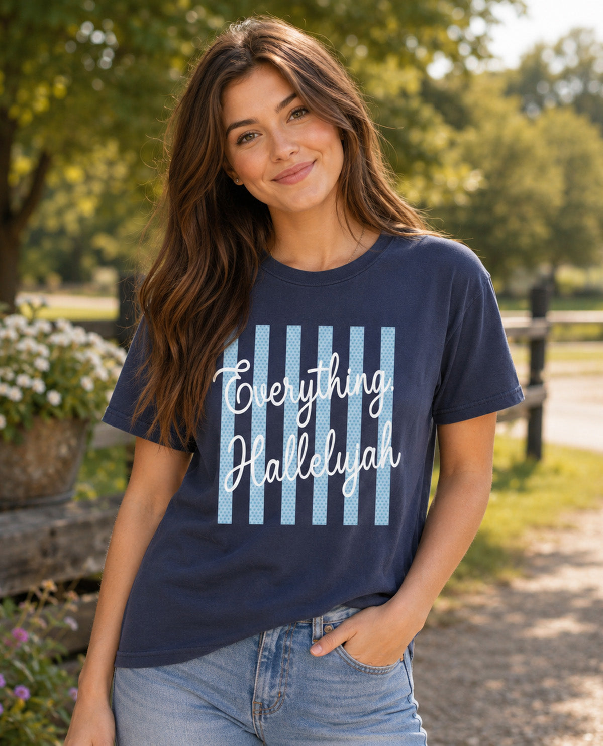 A woman wearing a navy blue graphic t-shirt with light blue vertical stripes and the text 'Everything Hallelujah' in white script, standing outdoors on a sunny path with trees and a fence in the background.