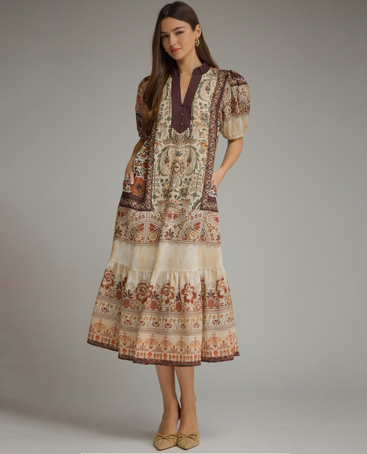 A woman models a tiered midi dress with a bohemian-style multicolor print in beige, brown, and rust tones, featuring puff sleeves and a deep V-neckline, set against a plain gray studio background.