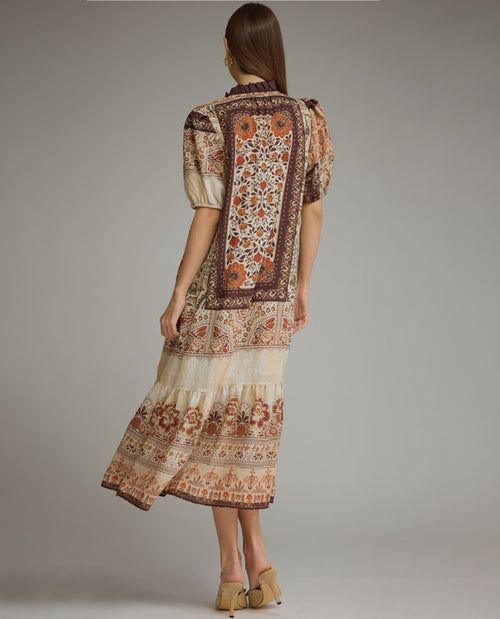A woman is shown from the back wearing a tiered midi dress with a brown and beige bohemian-style floral pattern, paired with tan pointed-toe flats, against a solid gray studio background.