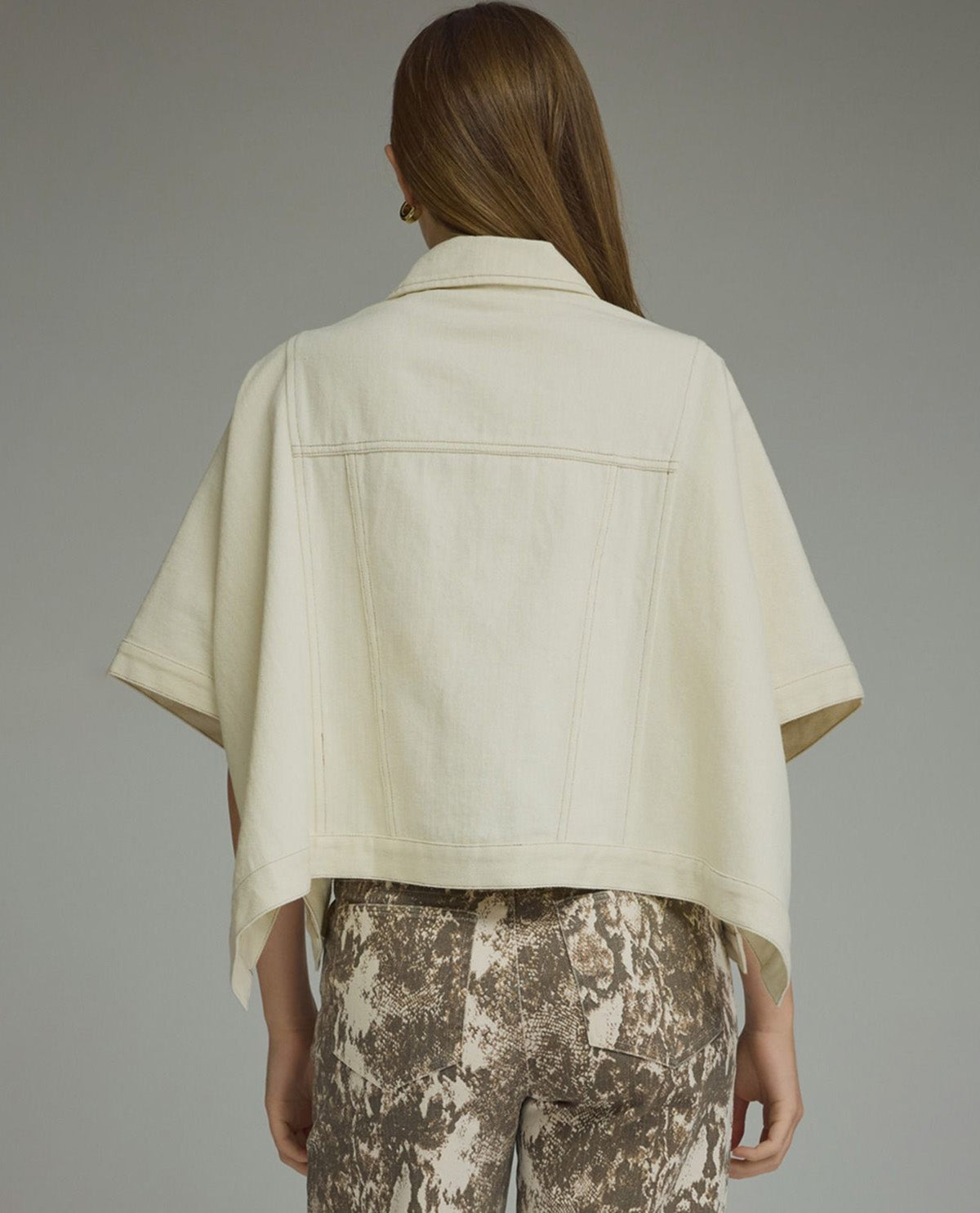 A model shown from the back wearing a cream-colored short-sleeve cropped jacket with a collar and a structured back panel, paired with brown and beige patterned pants, against a solid gray studio background.