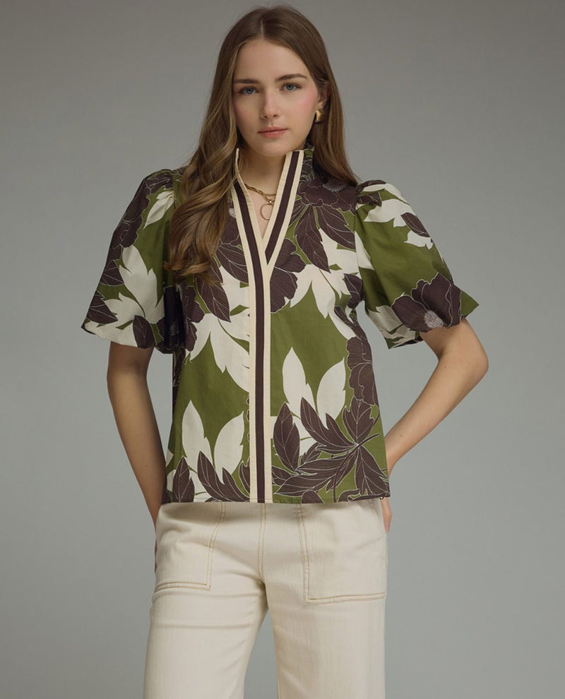 A woman modeling a short-sleeve button-front blouse with an olive green, cream, and dark brown floral pattern, paired with cream pants, against a solid gray studio background.