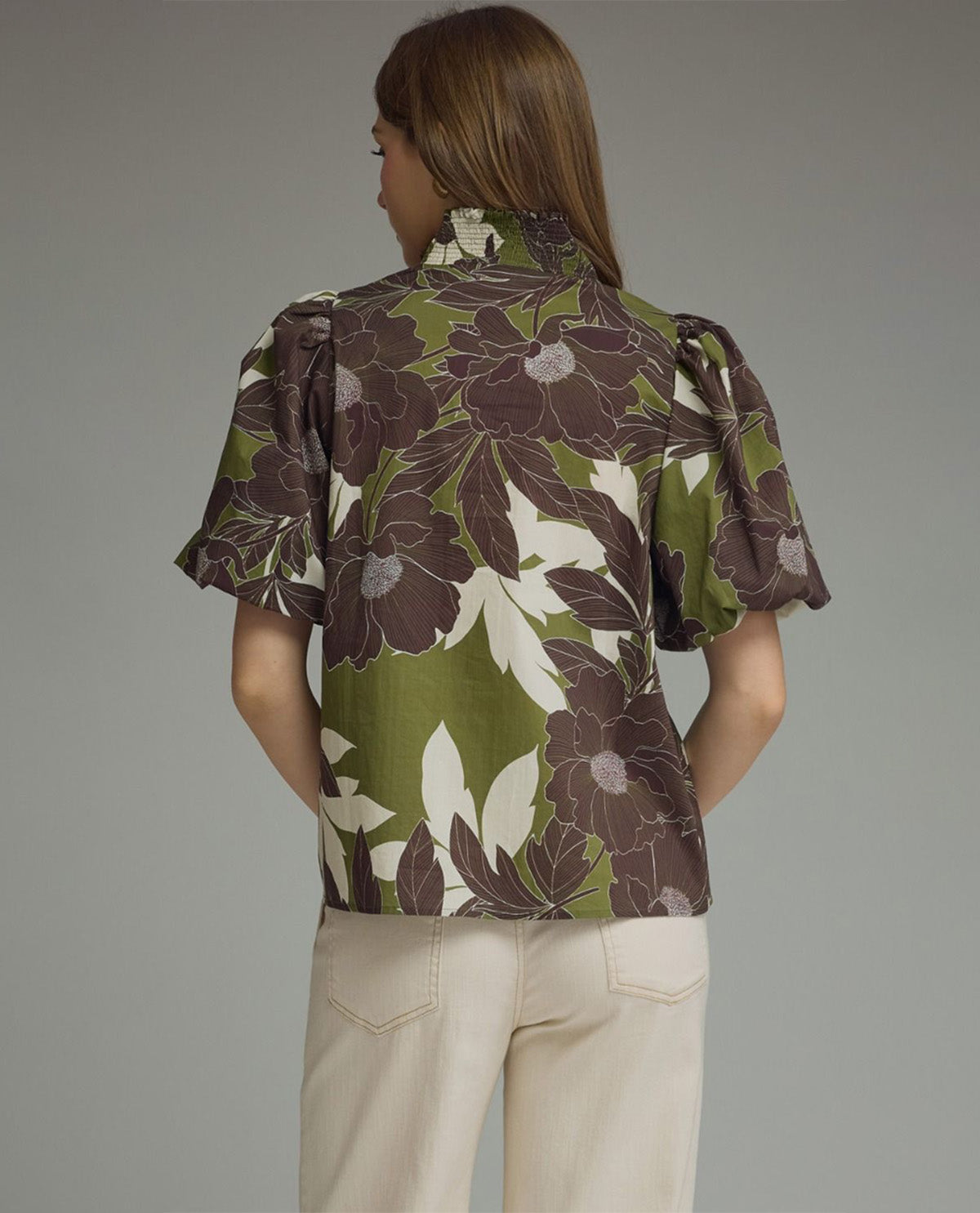 A woman viewed from the back wearing a short-sleeve floral blouse with olive green, brown, and cream tones, paired with light beige pants, against a plain gray studio background.