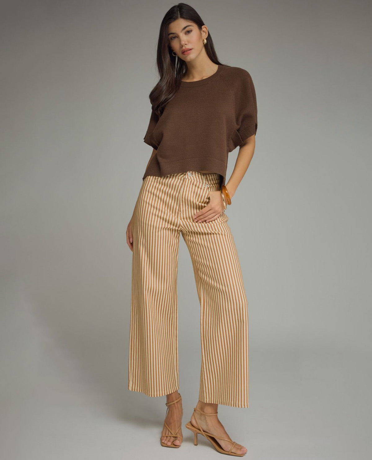 A model stands against a neutral gray studio background wearing a brown short-sleeve knit top, beige wide-leg cropped pants with thin vertical stripes, and nude high-heeled sandals.