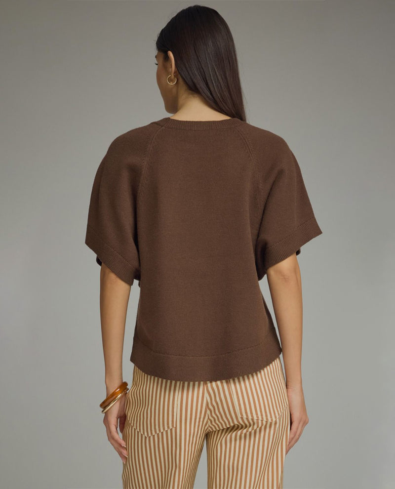 A woman shown from the back wearing a brown knit short-sleeve top and beige-and-white vertically striped trousers, photographed against a plain light gray studio background.