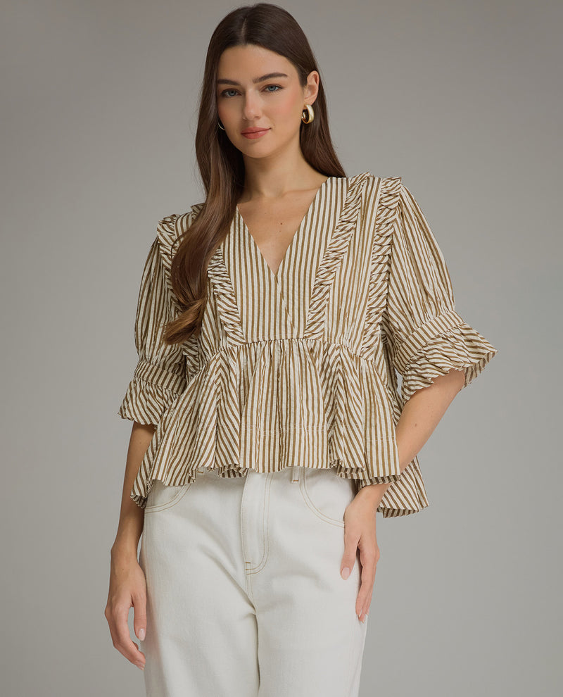 A woman modeling a beige and brown vertically striped blouse with ruffles and puff sleeves, paired with white pants, against a solid gray studio background.