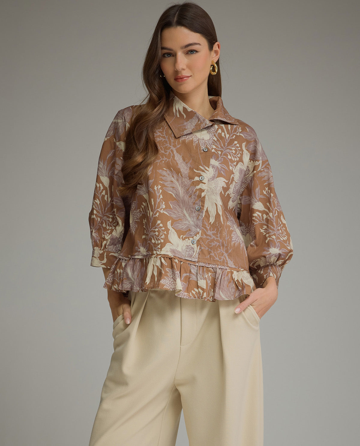 A woman modeling a brown and beige patterned blouse with a ruffled hem and wide sleeves, paired with light beige trousers, against a solid gray studio background.