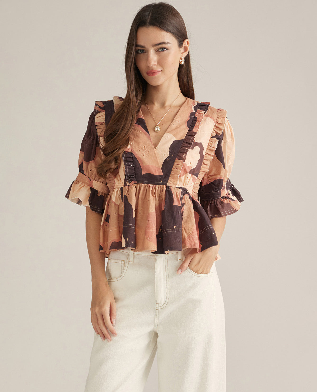 A woman modeling a short-sleeve ruffled blouse with a vintage-style art print in brown, beige, and black, paired with white pants, against a solid light gray background.