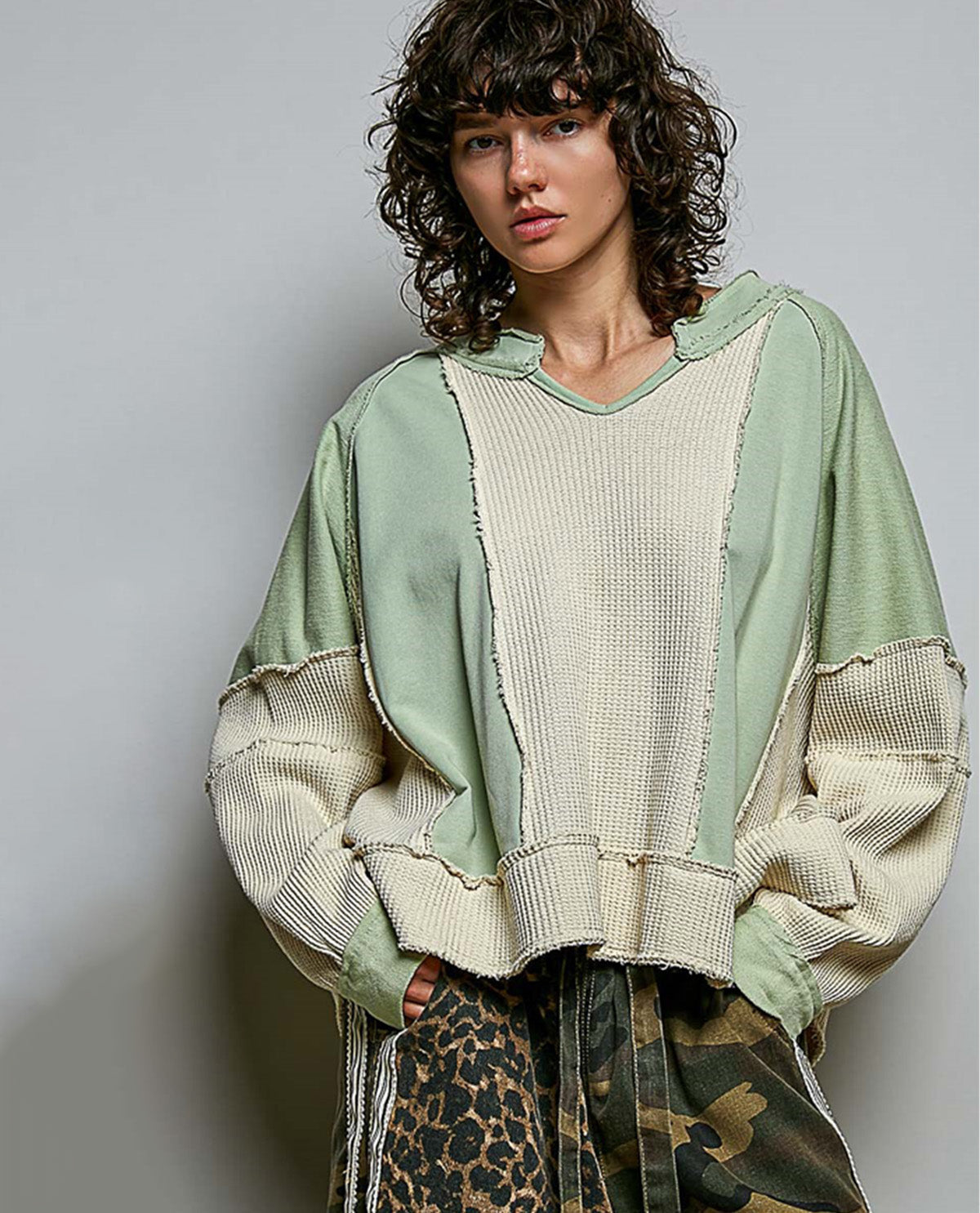 A model wearing a sage green and cream patchwork knit hoodie with raw-edge detailing, paired with a leopard print top and camouflage skirt, against a plain gray studio background.