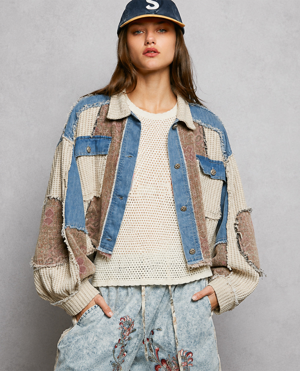 A model wearing a patchwork denim jacket with blue denim and beige textured knit panels, paired with a cream knit top and light blue jeans against a gray studio background.
