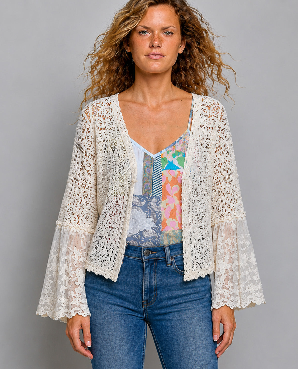 A woman modeling a cream lace cardigan with long bell sleeves over a colorful patterned camisole and blue jeans, against a solid gray studio background.