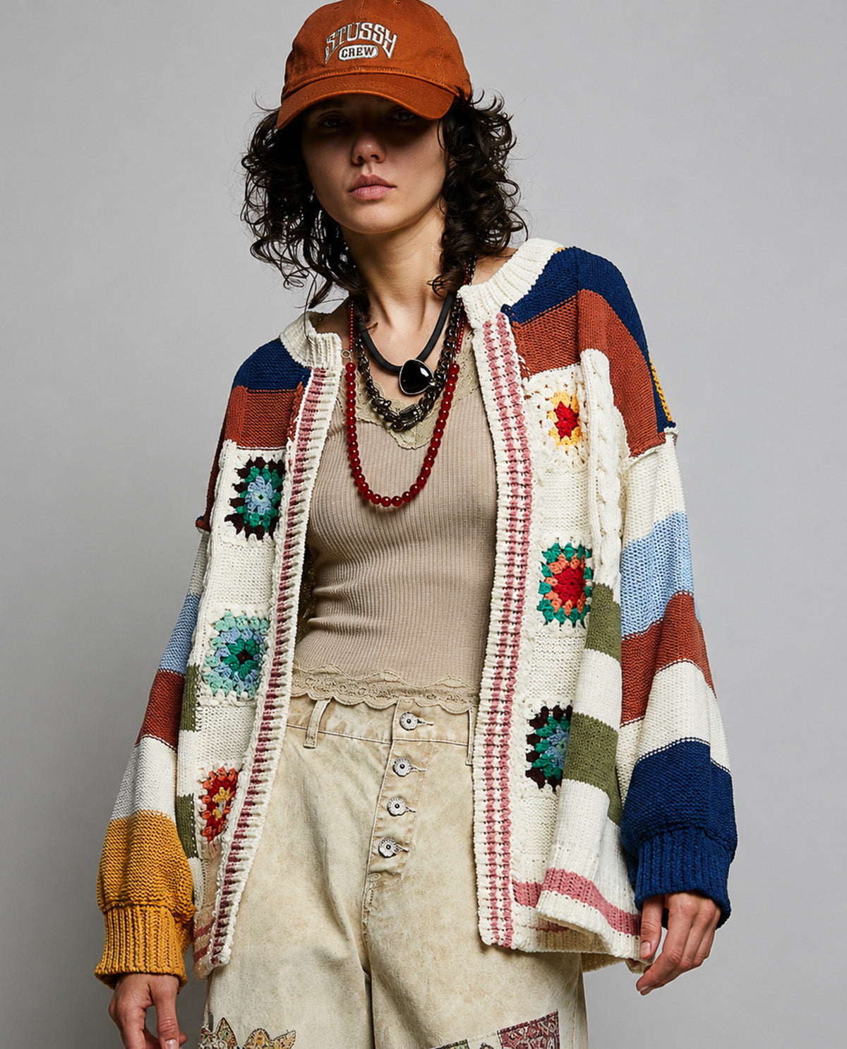 Model wearing a cream multi color cardigan with a tan shirt with beige bottoms an orange hat with a white background