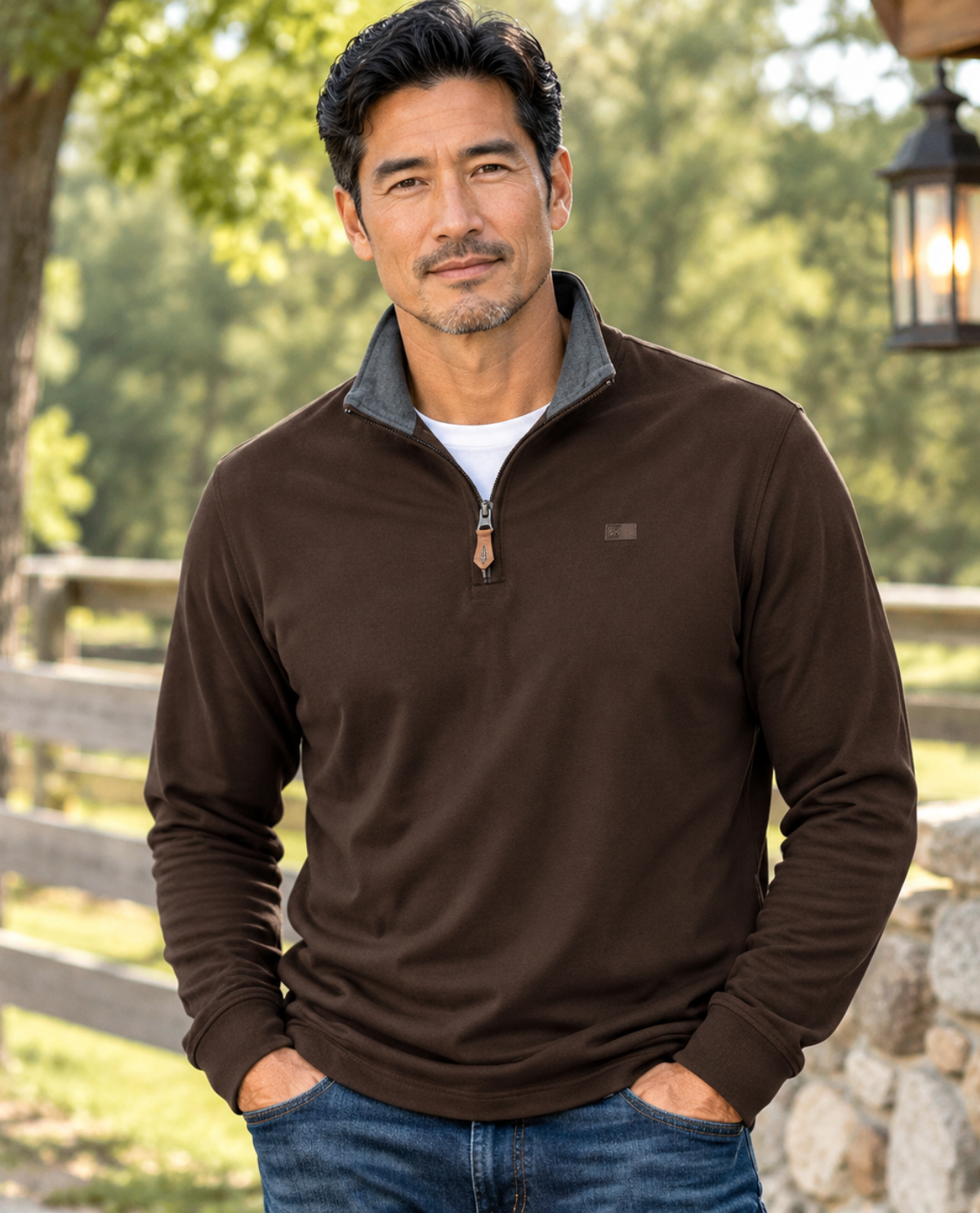 A man wearing a dark brown quarter-zip long-sleeve pullover with a gray collar, styled with blue jeans, standing outdoors in front of a rustic wooden fence and stone wall.