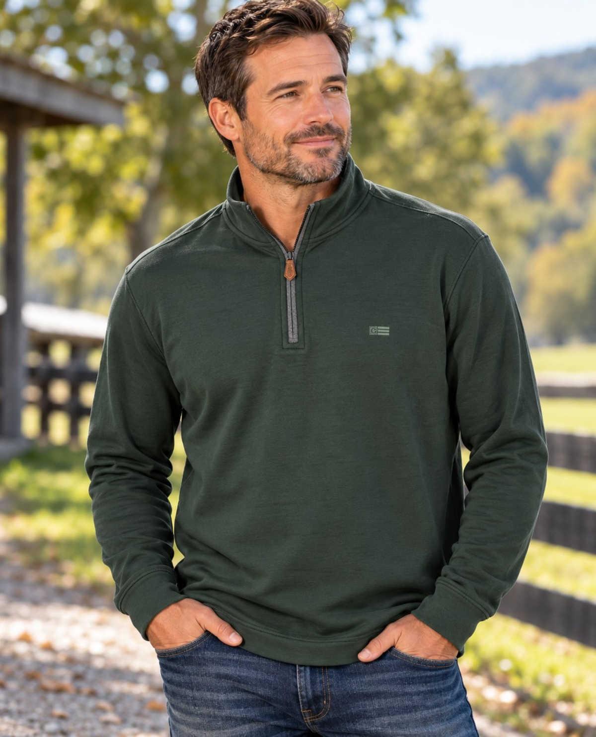 A man wearing a dark green quarter-zip long-sleeve pullover with blue jeans, standing outdoors in a rural setting with trees and a fence in the background.