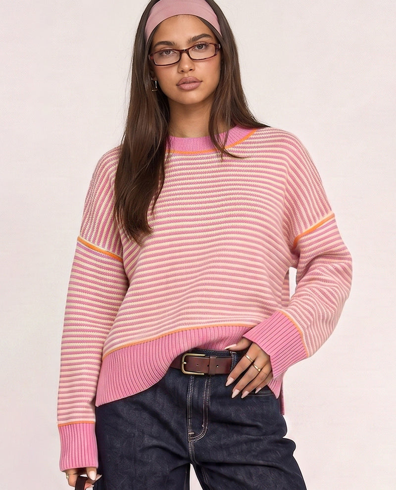 A woman wearing a pink and white striped knit sweater with orange-yellow stripe accents on the shoulders and cuffs, paired with dark blue jeans and a matching pink headband, against a light beige studio background.