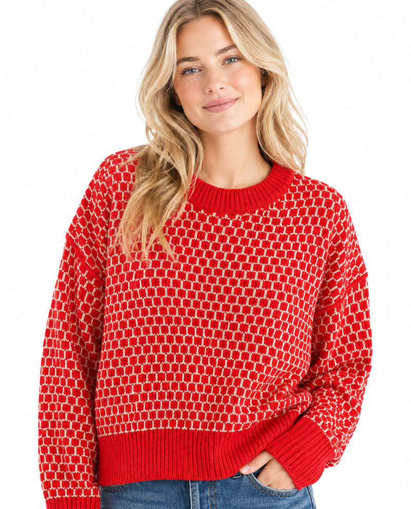 A woman wearing a bright red knit sweater with a white geometric pattern, paired with blue jeans, against a plain white background.