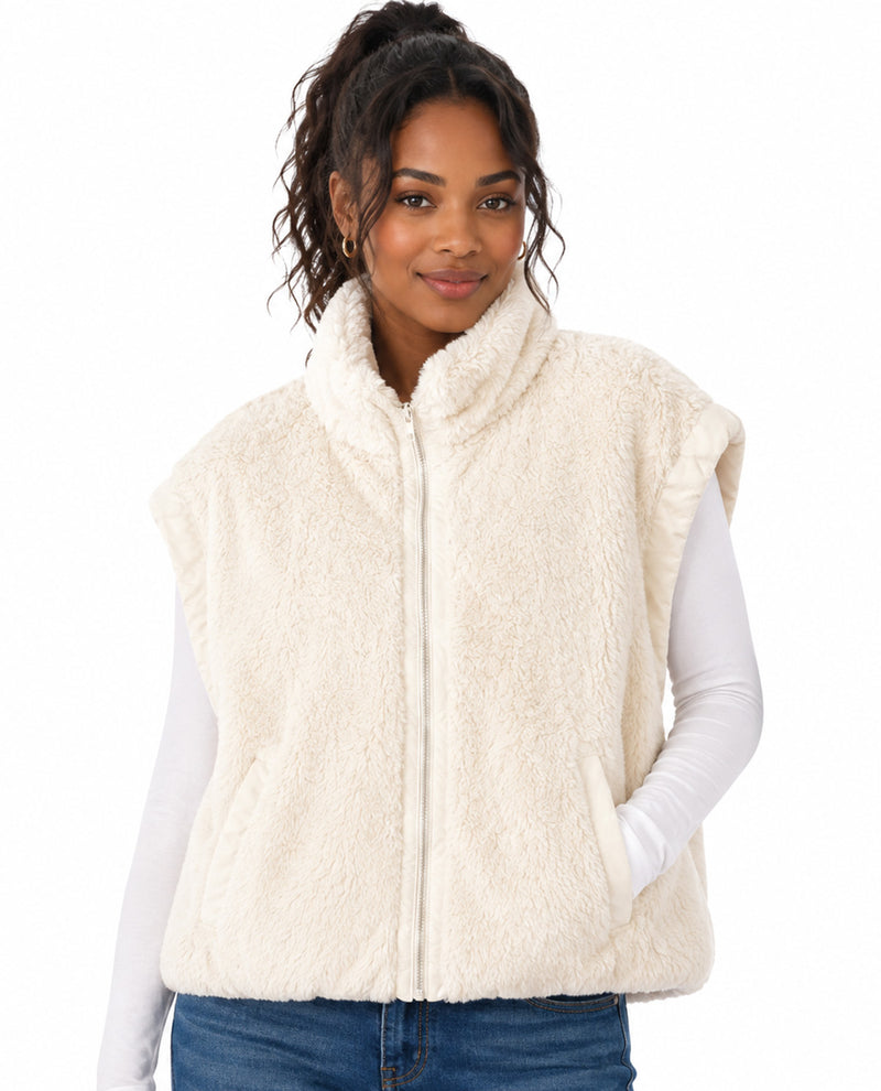 A woman wearing a cream-colored, plush faux-fur zip-up vest over a white long-sleeve shirt and blue jeans, photographed against a solid white background.