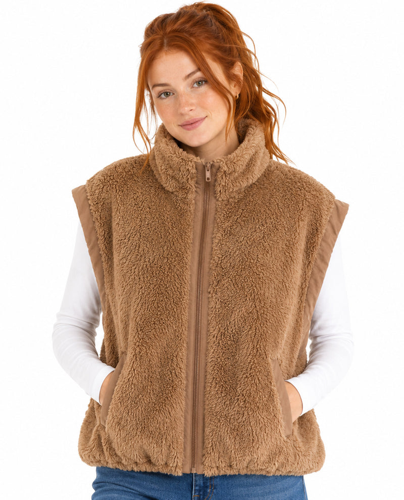 A woman with red hair wearing a brown sherpa fleece zip-up vest over a white long-sleeve shirt and blue jeans, photographed against a plain white background.