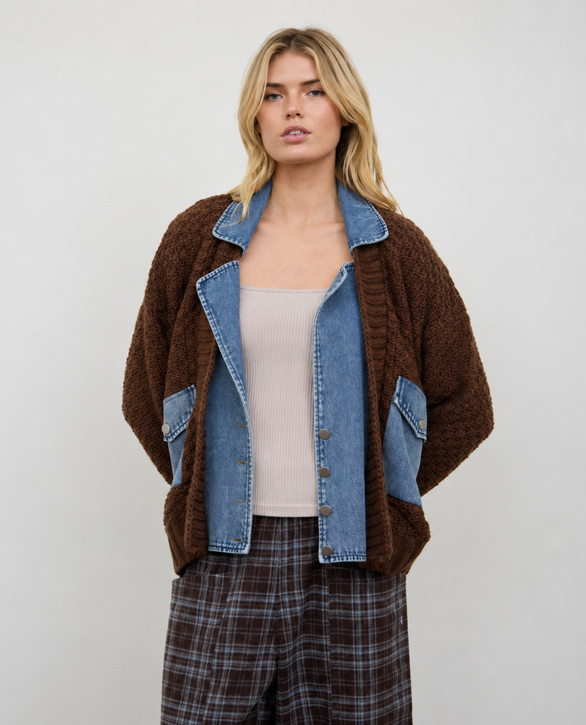 A woman modeling a brown fuzzy textured cardigan with light blue denim panels, worn over a beige ribbed tank top and brown plaid trousers, against a plain light gray studio background.