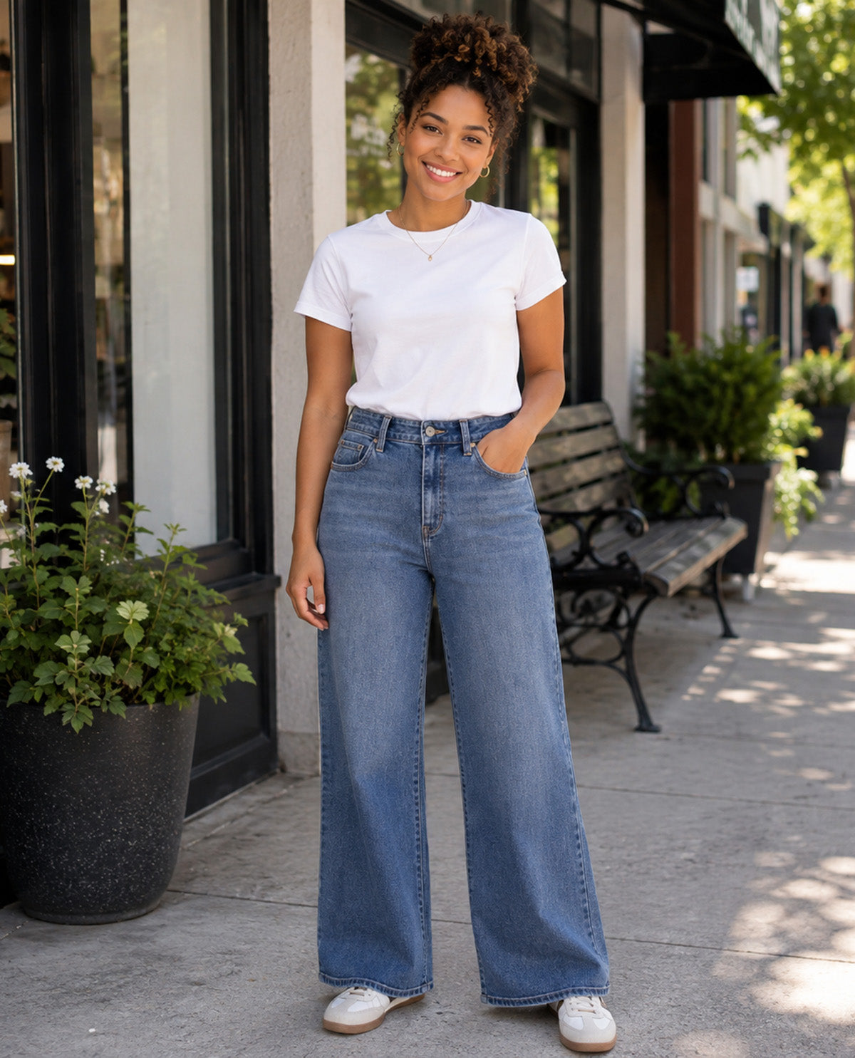A woman stands on a city sidewalk wearing a white short-sleeve crewneck t-shirt and high-waisted, wide-leg blue jeans with white sneakers. The background shows storefront windows, potted plants, and a wooden bench.