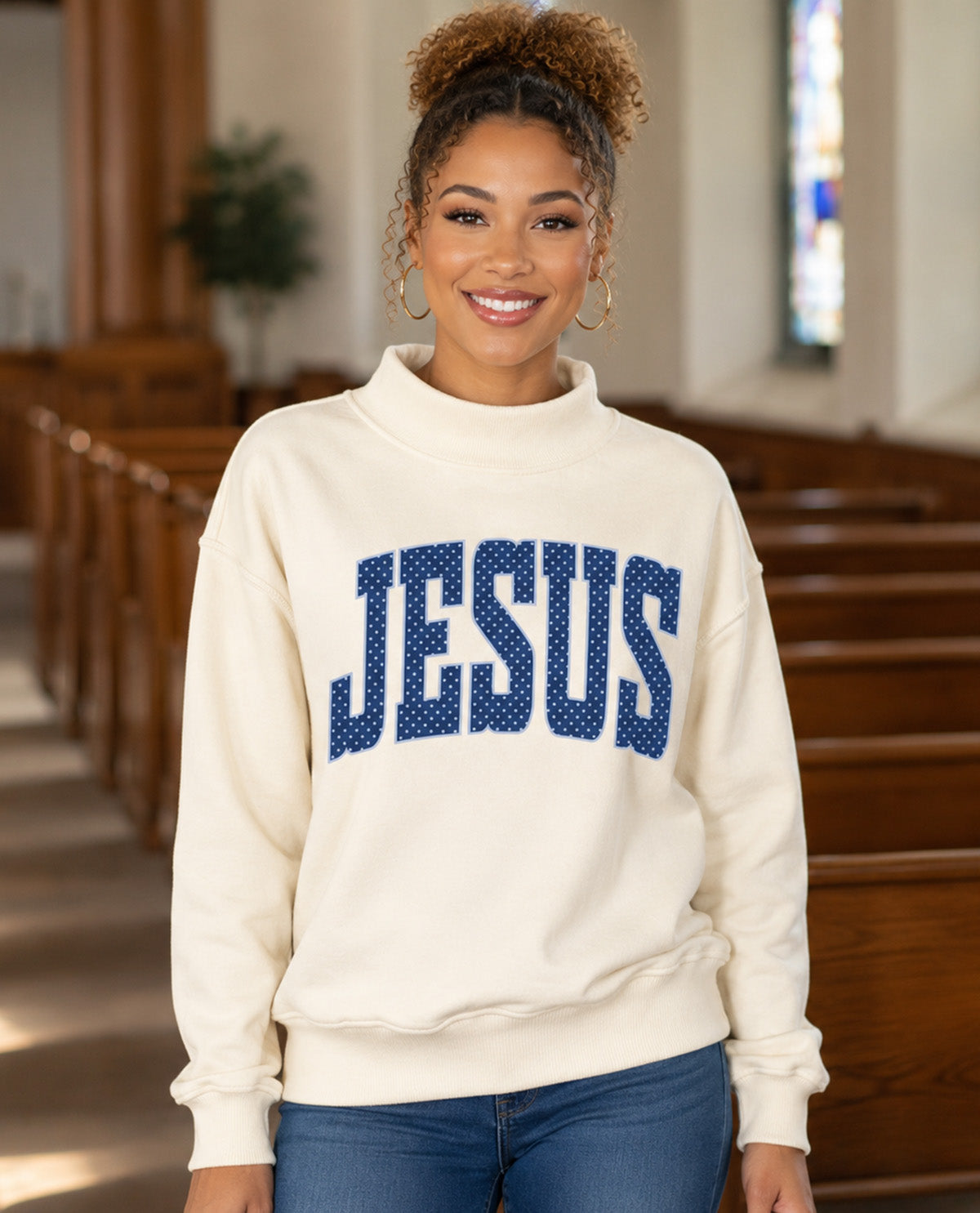A woman wearing a cream-colored mock neck sweatshirt with 'JESUS' in blue dotted varsity letters, standing in a church interior with wooden pews in the background.