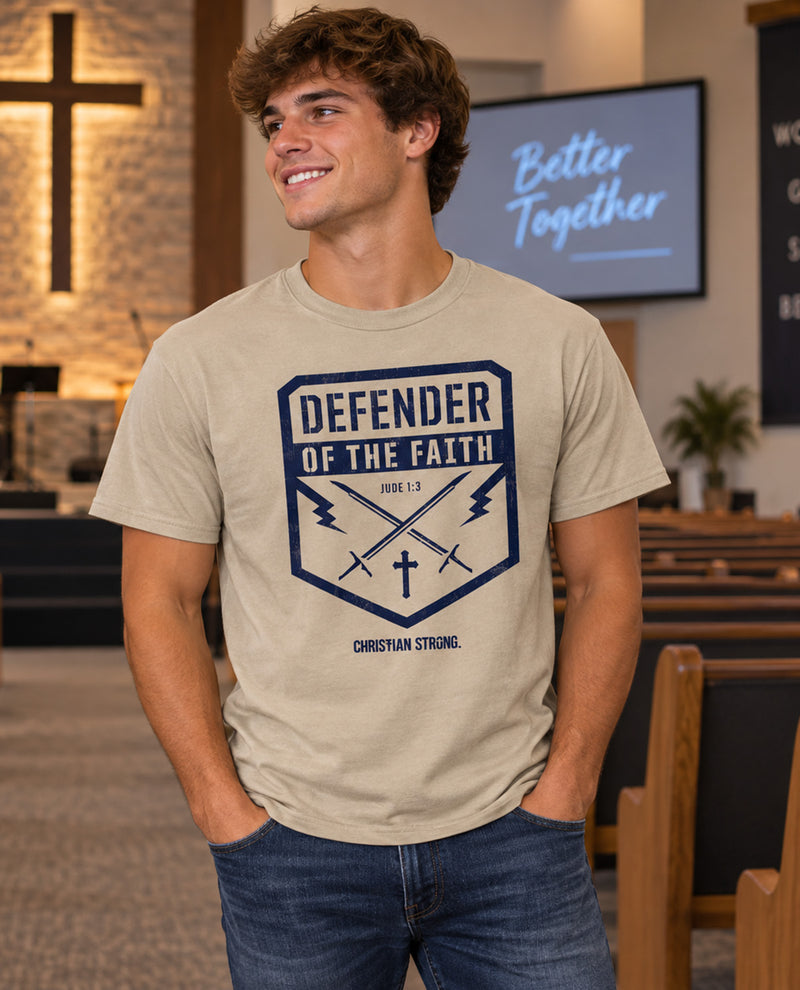 A man wearing a beige graphic t-shirt with a navy blue 'Defender of the Faith' design, standing inside a church with pews and a screen in the background.