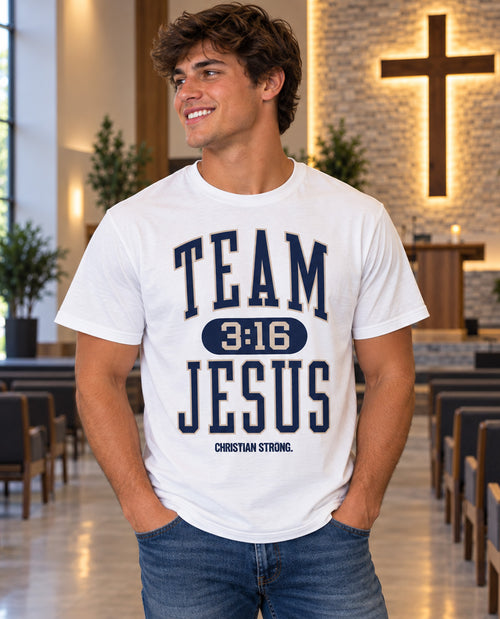 A smiling young man wearing a white graphic t-shirt with 'TEAM JESUS 3:16 CHRISTIAN STRONG.' printed in navy blue, standing inside a church hall with pews and a cross on the wall in the background.