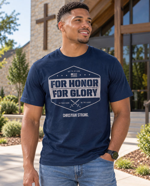 A man wearing a navy blue graphic t-shirt with the text 'FOR HONOR FOR GLORY' and 'CHRISTIAN STRONG.' printed on the front, standing outdoors in front of a modern church building.