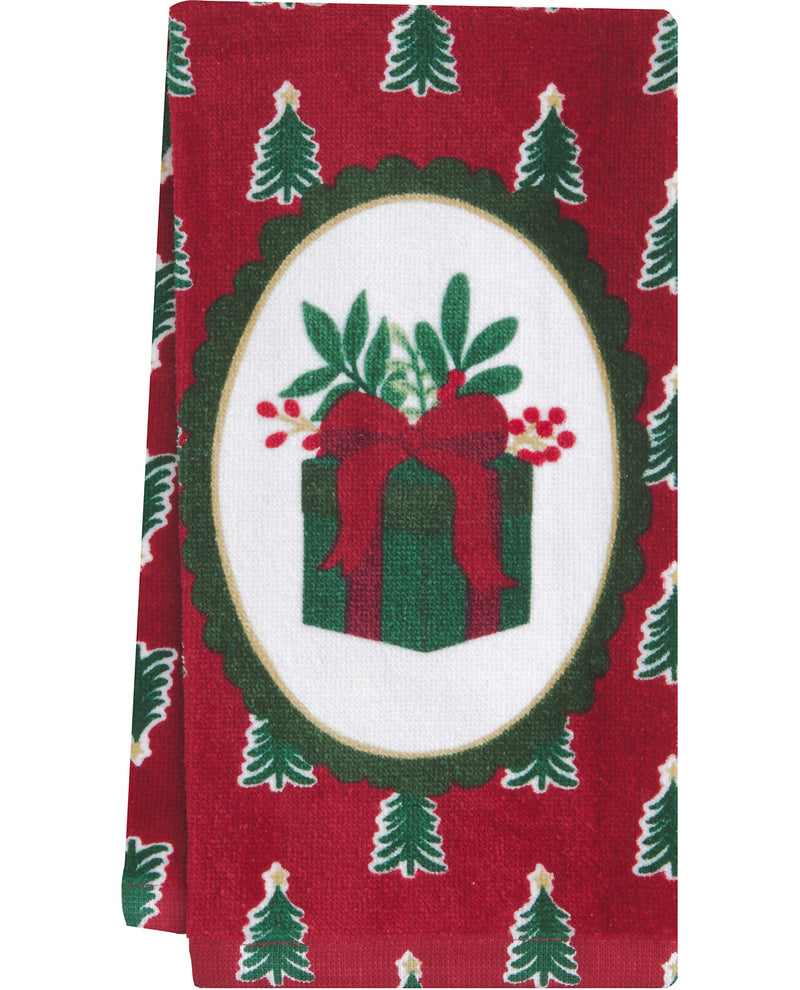A red holiday-themed fabric napkin or tea towel featuring a central white oval with a green gift box, red bow, green leaves, and red berries, surrounded by a green scalloped border and small green Christmas trees on a red background.