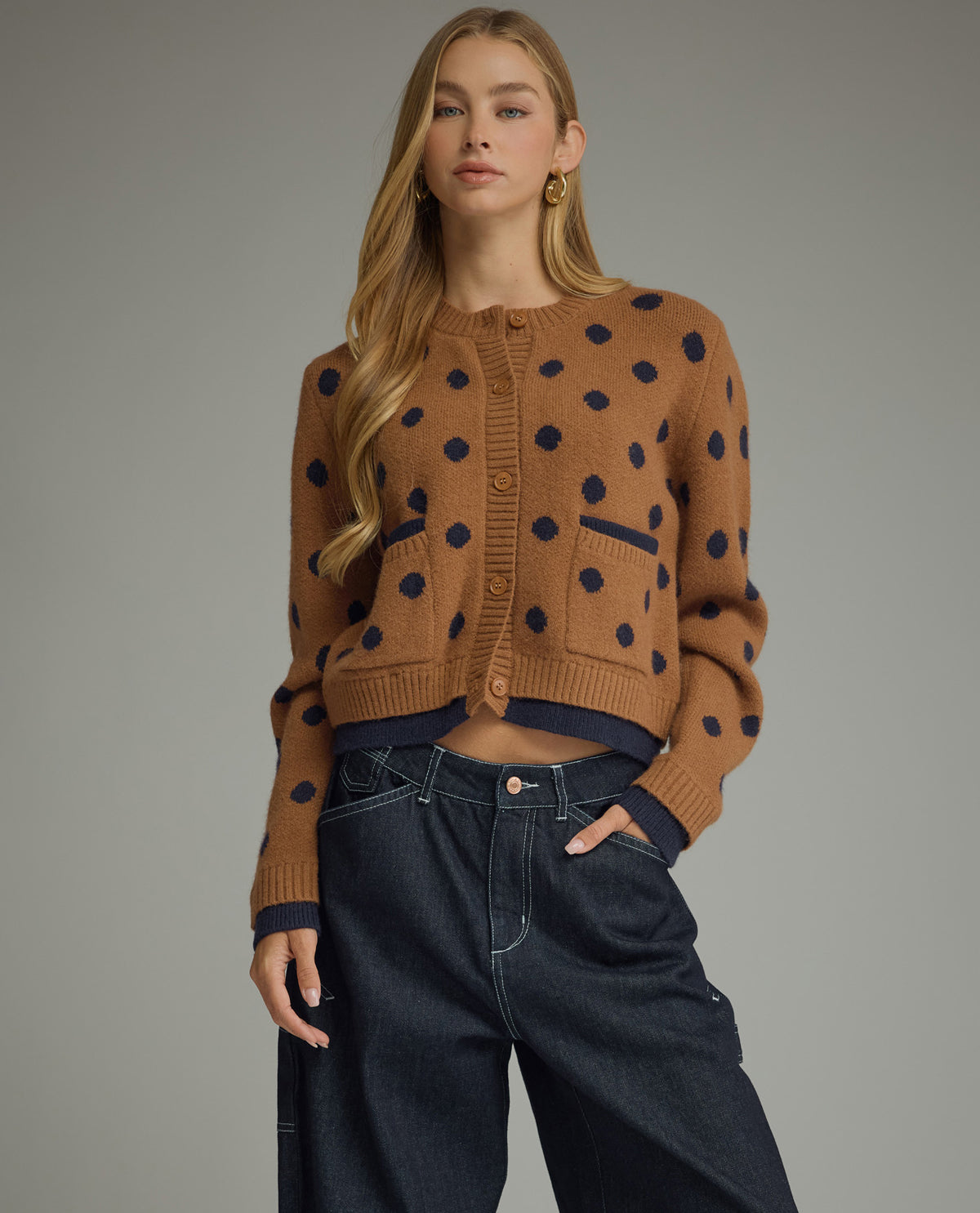 A woman modeling a cropped brown knit cardigan with dark navy blue polka dots, paired with dark wide-leg jeans, against a solid gray studio background.