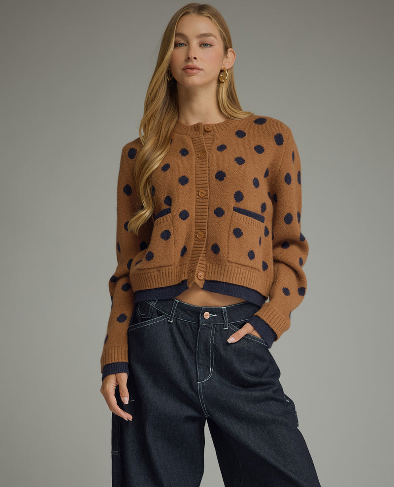 A woman modeling a cropped brown knit cardigan with dark navy blue polka dots, paired with dark wide-leg jeans, against a solid gray studio background.