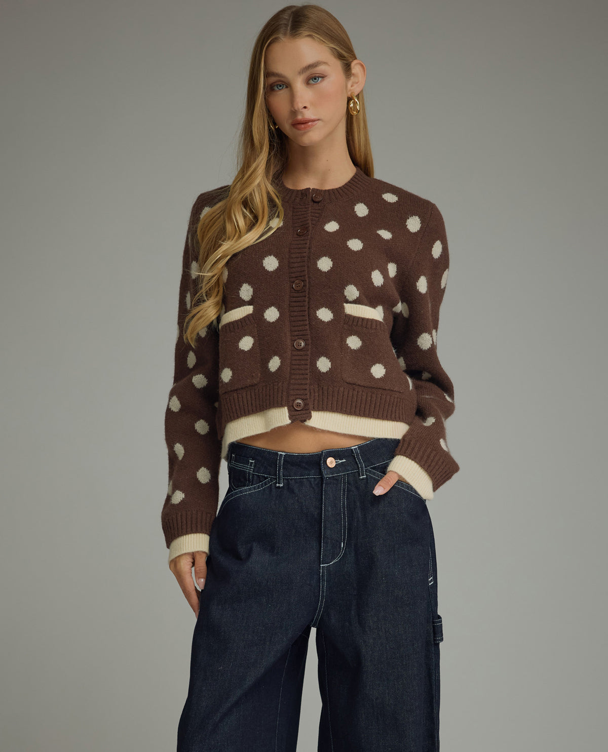 A woman modeling a brown cropped knit cardigan with cream polka dots and ribbed trim, paired with dark blue jeans, against a solid gray studio background.