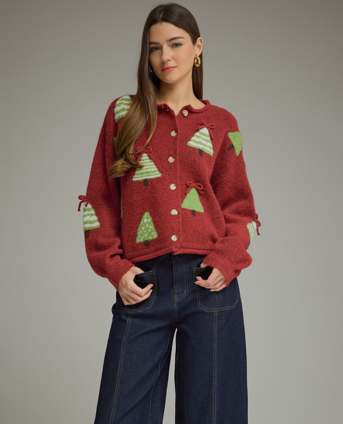 A woman modeling a red fuzzy knit cardigan with green Christmas tree patterns, paired with dark blue jeans, against a solid gray studio background.