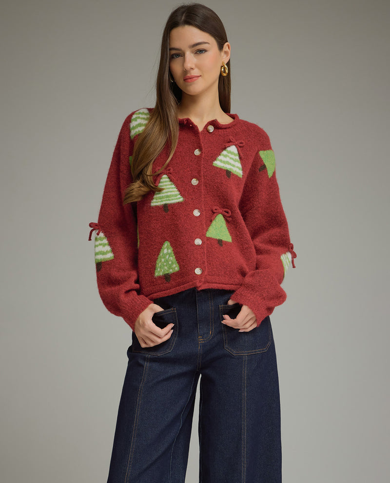 A woman modeling a red fuzzy knit cardigan with green Christmas tree patterns, paired with dark blue jeans, against a solid gray studio background.