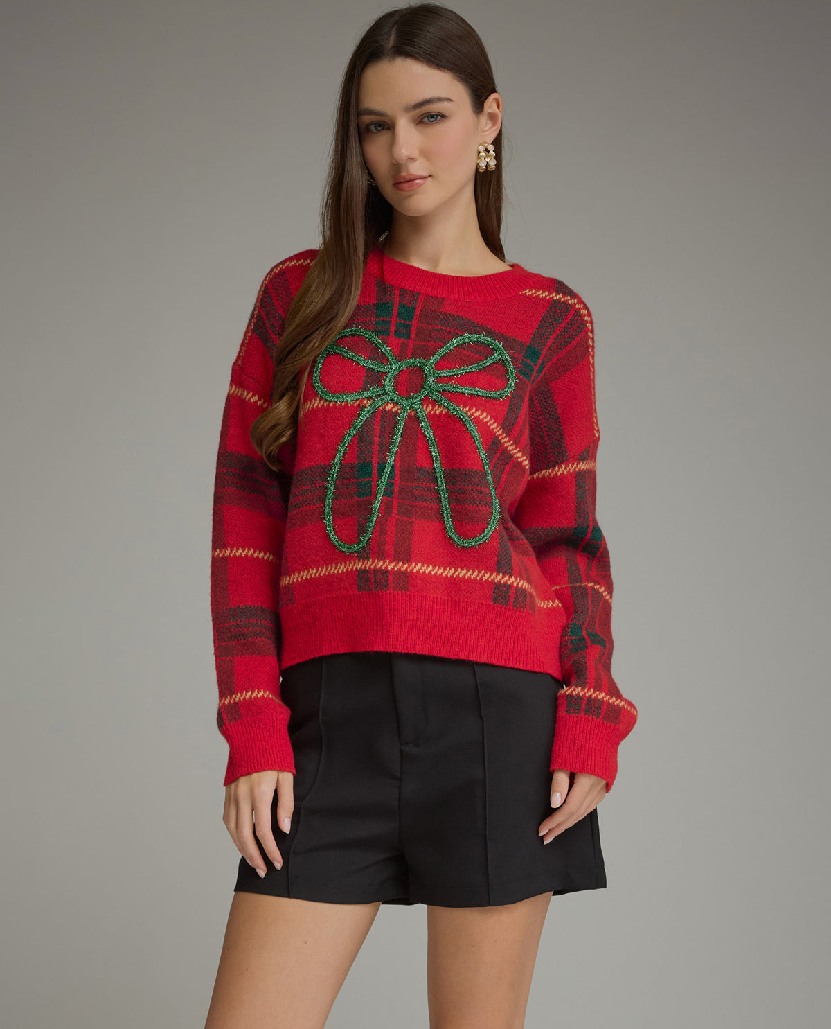A woman modeling a red knit sweater with a green beaded bow motif on the front, paired with black shorts, against a solid gray studio background.