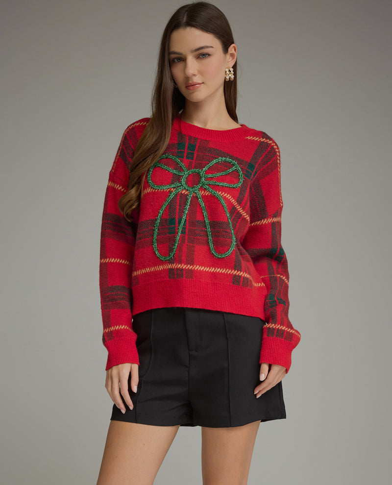 A woman modeling a red knit sweater with a green beaded bow motif on the front, paired with black shorts, against a solid gray studio background.