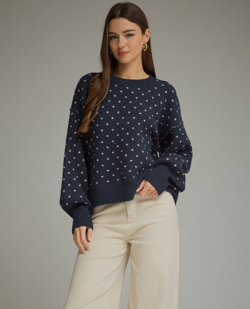 A woman modeling a navy blue crewneck sweater with a white star pattern, paired with cream-colored pants, against a solid gray studio background.
