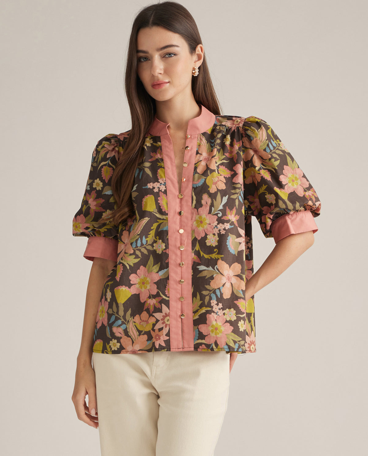 A woman modeling a short-sleeve button-front blouse with a dark brown floral print, pink collar, cuffs, and placket, paired with cream pants against a plain beige studio background.