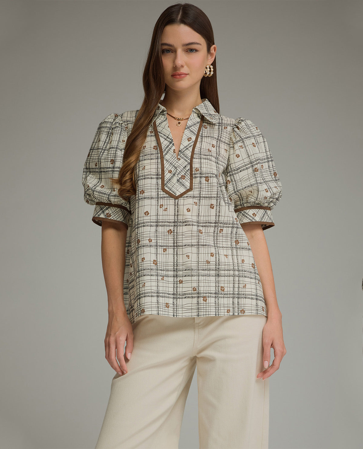 A woman modeling a short-sleeve plaid blouse with puff sleeves and a decorative V-neckline, paired with light beige trousers, against a solid gray studio background.