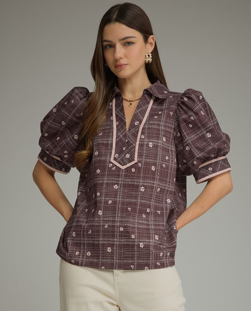 A woman modeling a short-sleeve button-up blouse with puff sleeves, featuring a dark plum purple and pink plaid pattern with small white floral accents, paired with cream pants against a solid gray studio background.