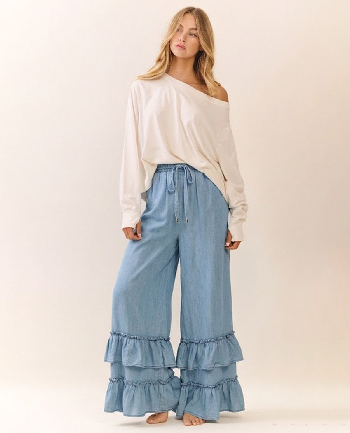 A woman modeling a cream off-the-shoulder long-sleeve top and light blue wide-leg ruffled pants against a plain beige studio background.