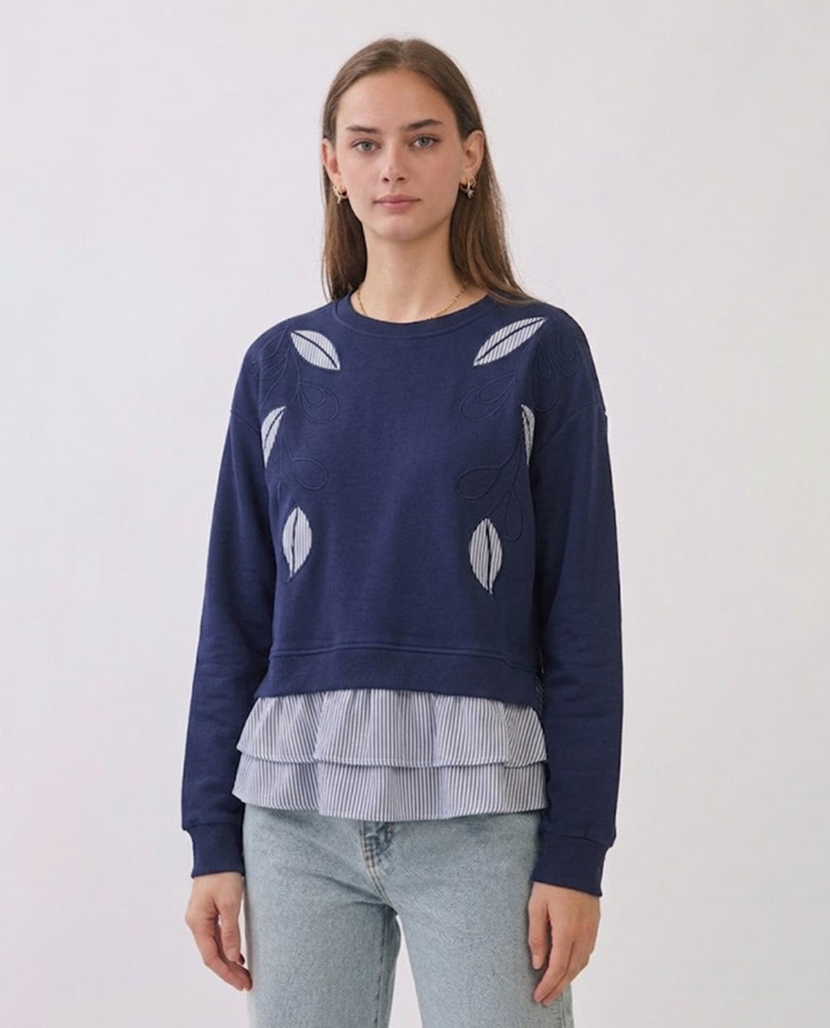 A woman modeling a navy blue crewneck sweatshirt with white leaf embroidery and a layered ruffled light blue and white striped hem, paired with light-wash jeans against a plain white studio background.