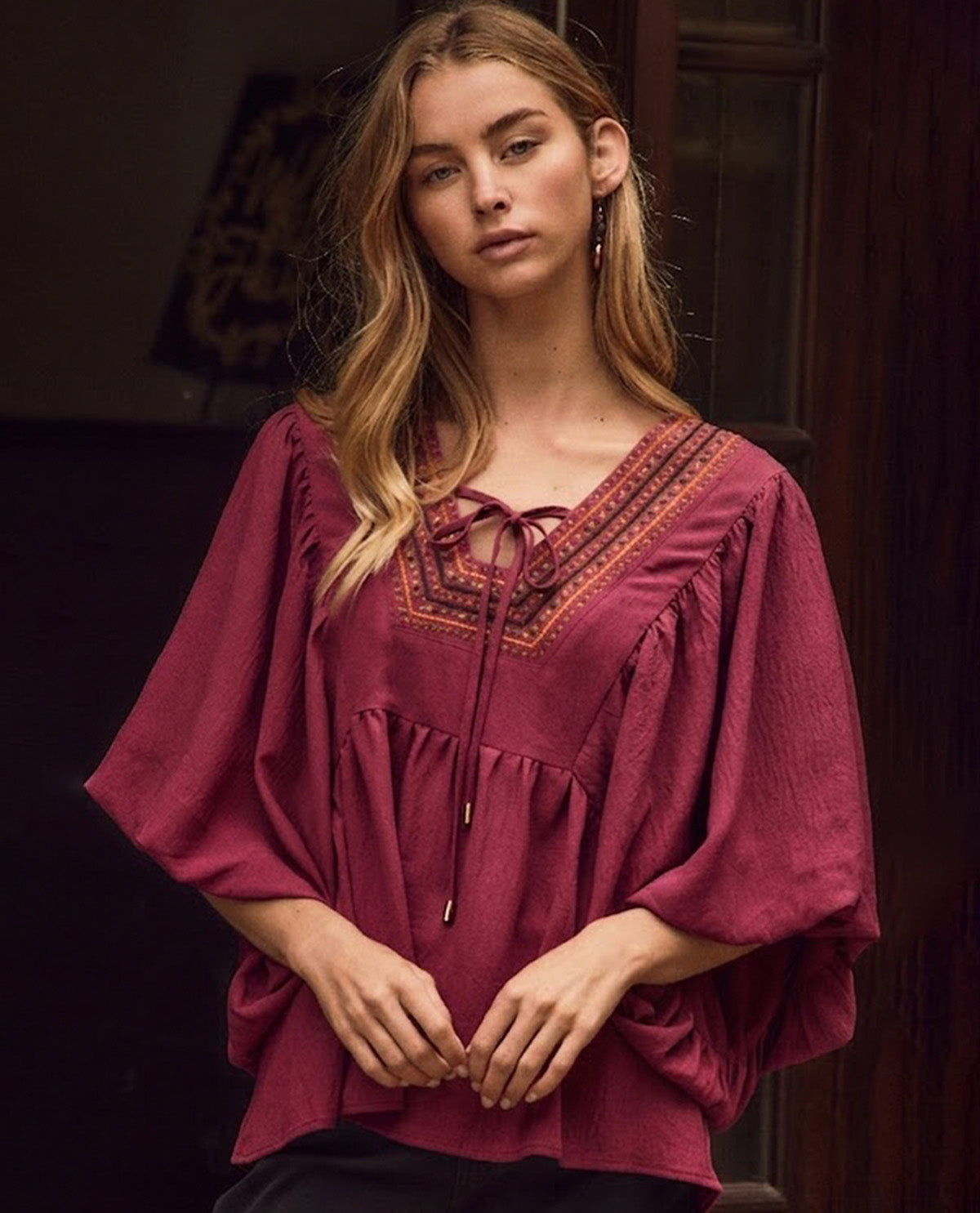 A woman wearing a loose, deep burgundy blouse with wide three-quarter sleeves, a V-neckline, and embroidered detailing around the neckline with a front tie closure, posed against a dark, rustic background.