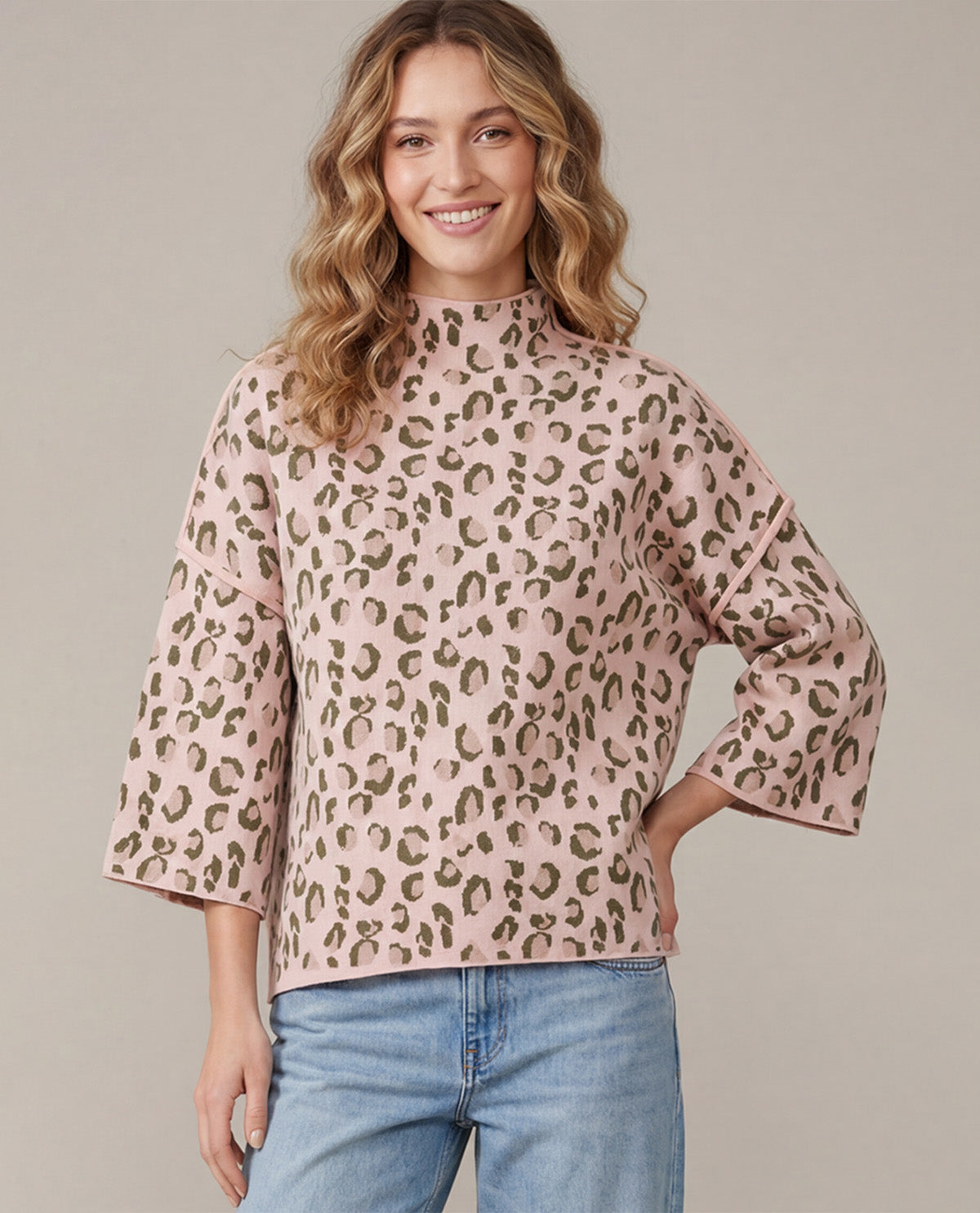 A woman wearing a pink leopard-print mock neck top with three-quarter bell sleeves and light blue jeans, standing against a solid beige studio background.