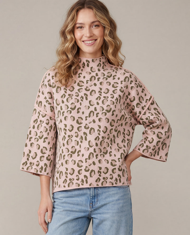 A woman wearing a pink leopard-print mock neck top with three-quarter bell sleeves and light blue jeans, standing against a solid beige studio background.