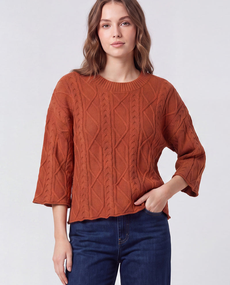 A woman wearing a rust-orange cable-knit sweater with three-quarter sleeves and dark blue jeans, standing against a light gray studio background.