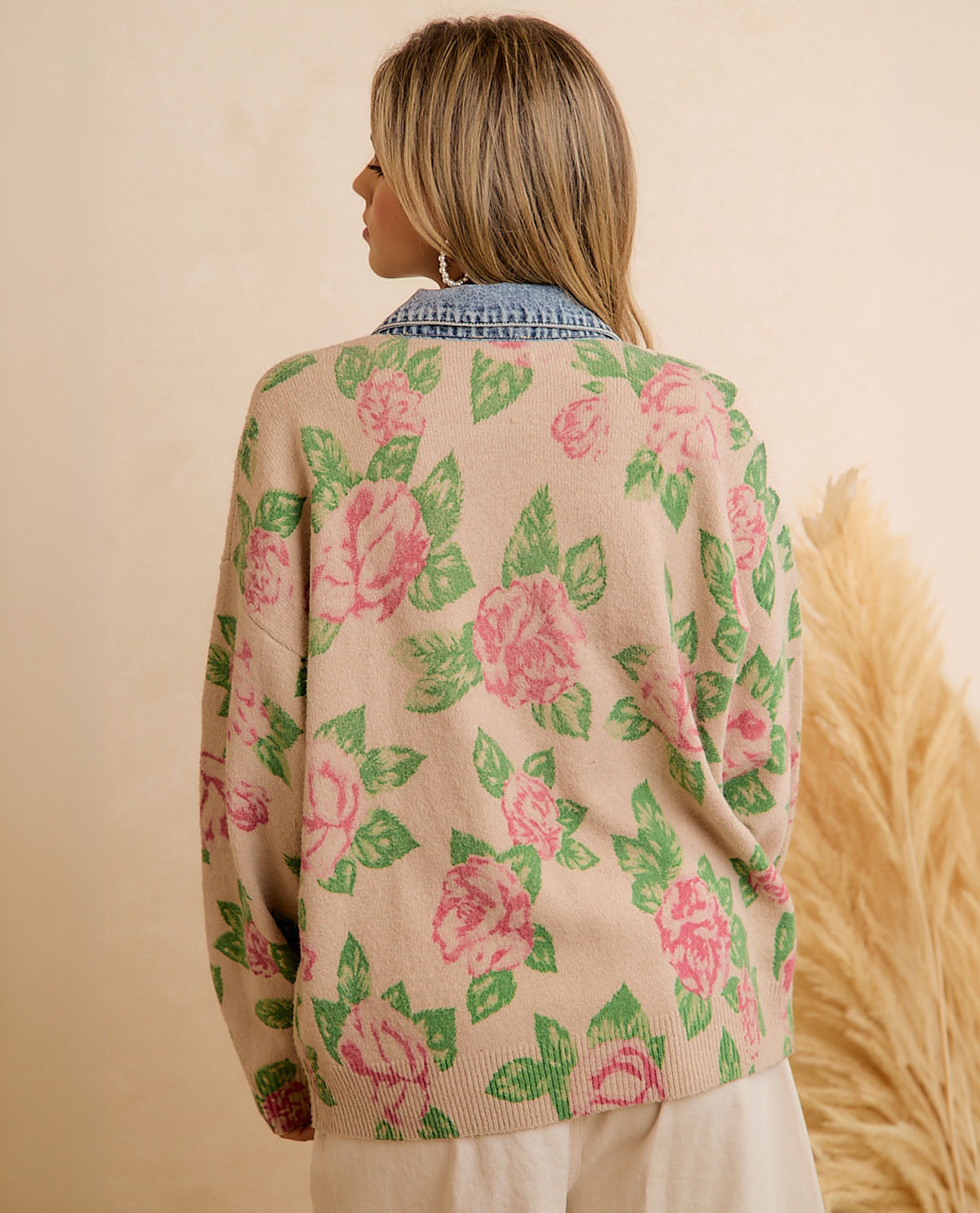A woman shown from the back wearing a beige knit cardigan with a pink rose and green leaf pattern, layered over a light blue denim shirt. The background is a warm beige wall with a dried pampas grass decoration on the right.