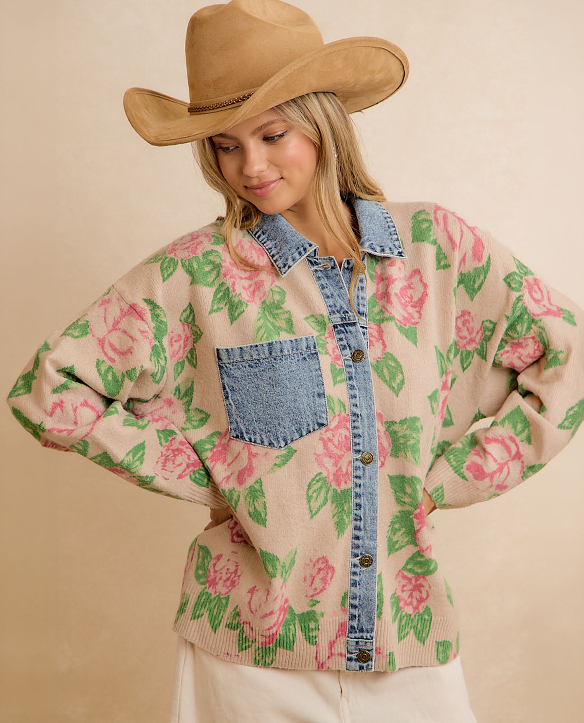 A woman wearing a beige cardigan with a pink rose and green leaf floral pattern, featuring a blue denim collar, button placket, and chest pocket, paired with a tan cowboy hat against a beige studio background.
