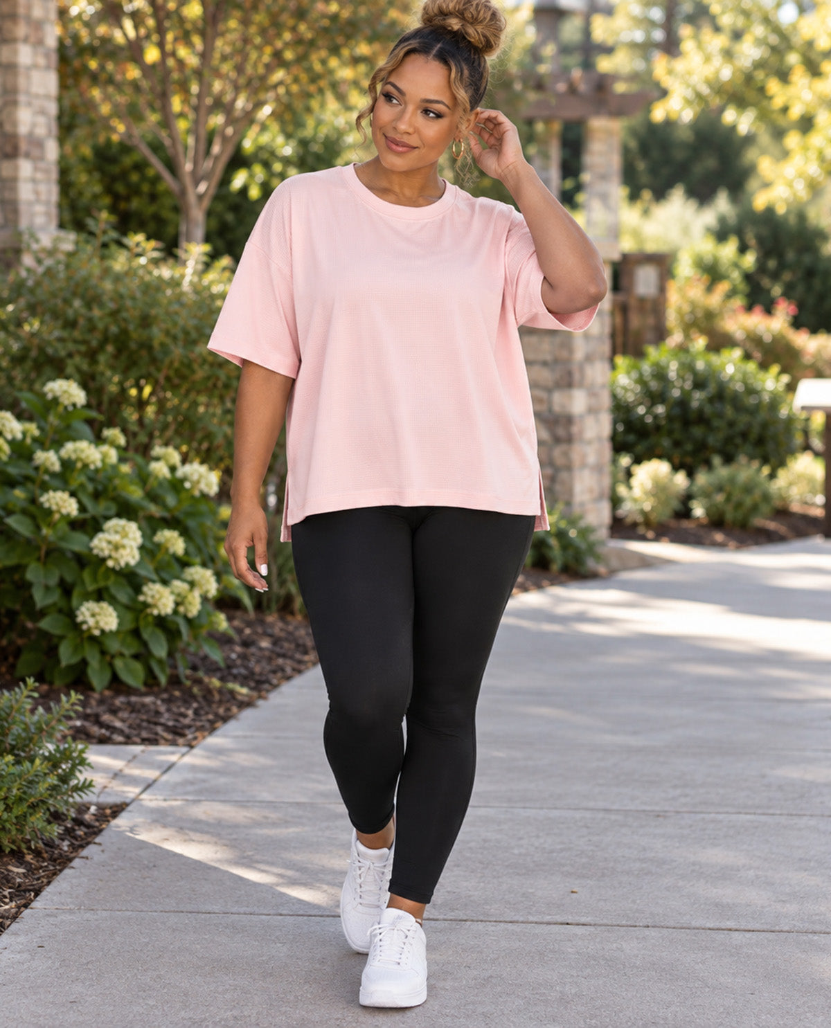 A woman walking outdoors on a paved path wearing a light pink oversized short-sleeve t-shirt, black leggings, and white sneakers.